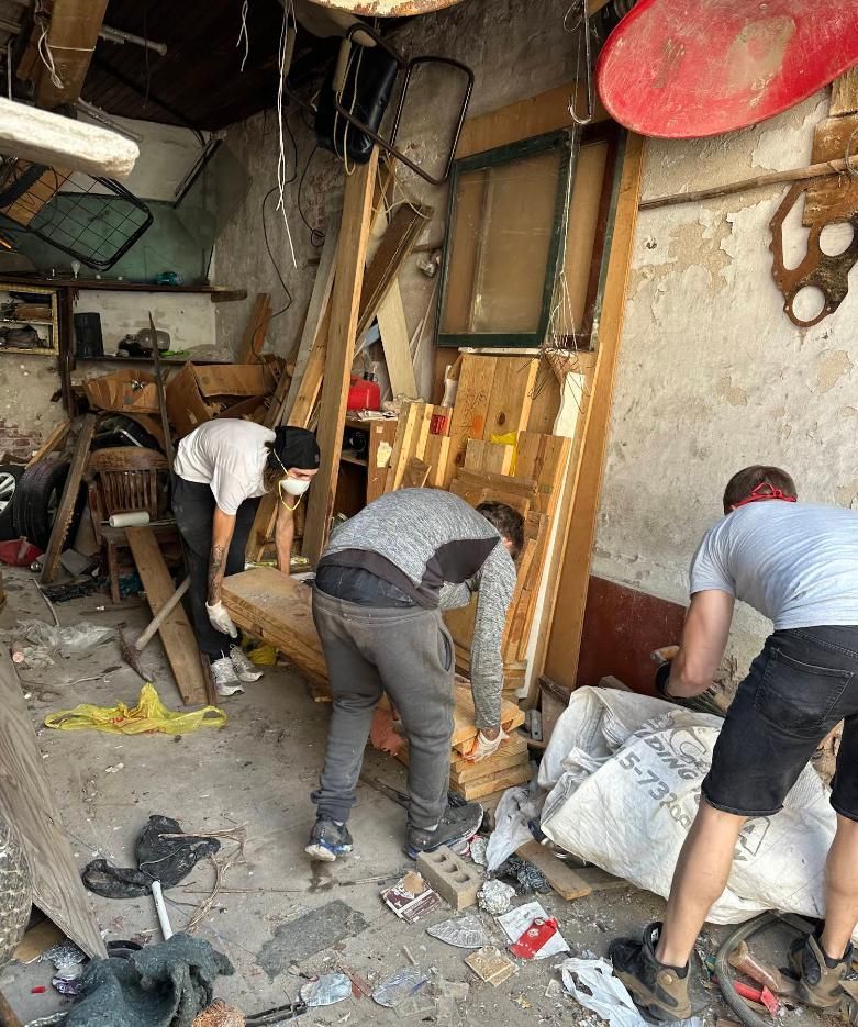Three people clearing debris in a cluttered workshop or garage with broken items and scattered rubble.