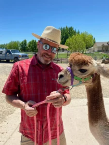 A man in a hat is holding a leash next to an alpaca.