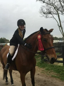 A woman is riding a brown horse with a red ribbon around its neck