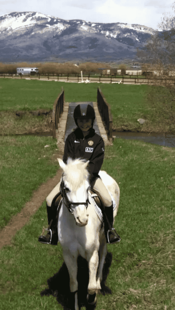 A person is riding a white horse in a field with mountains in the background.