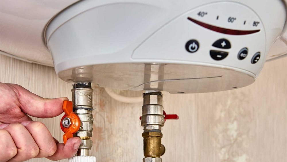Hand turning an orange valve on a water heater; white appliance, light background.