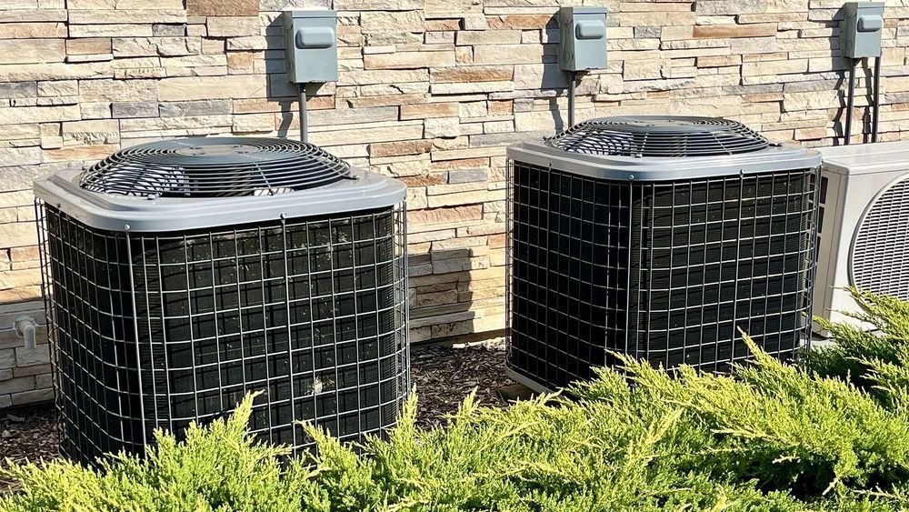 Two air conditioning units outside a building, with a stone facade and shrubs.