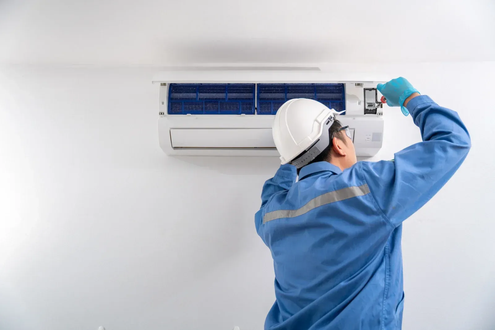 HVAC technician in blue uniform, white helmet, gloves, working on an air conditioning unit on a white wall.