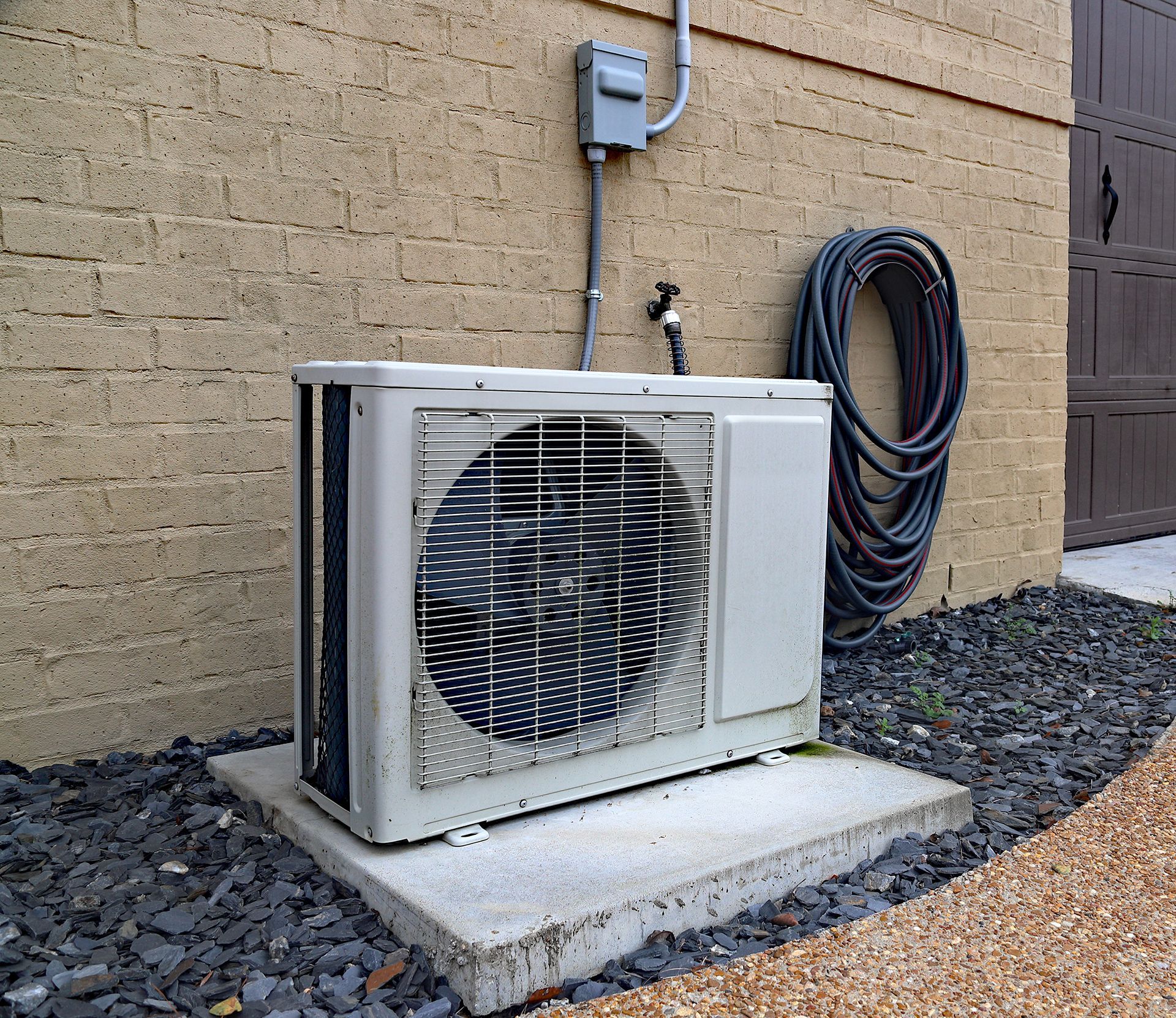 A white air conditioner is sitting outside of a brick building next to a hose.