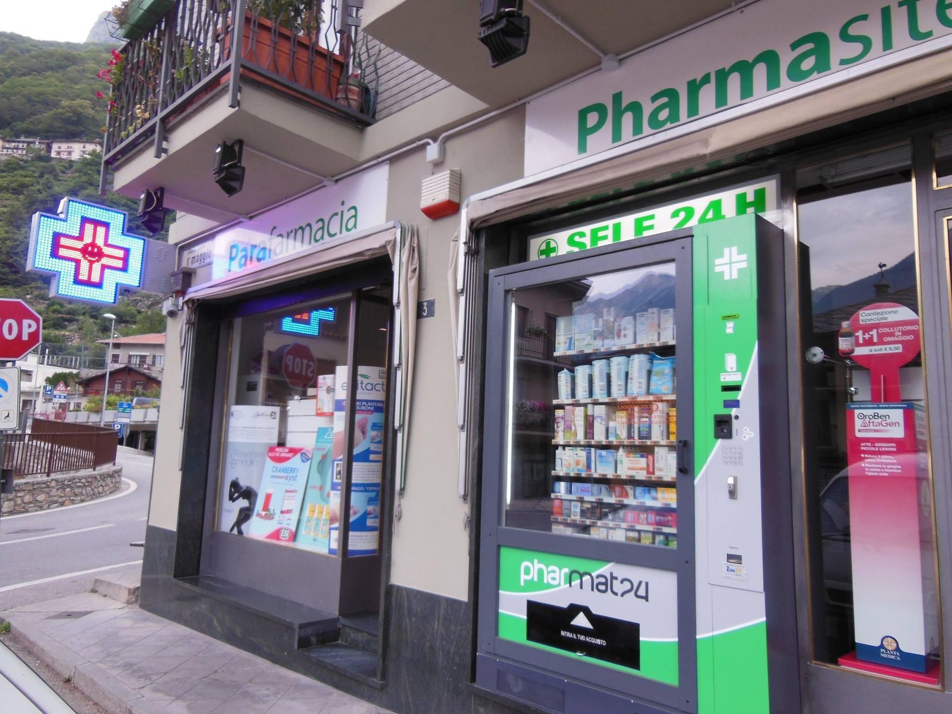 La vetrina di una farmacia con un'insegna al neon a forma di croce e, all'esterno, un distributore automatico verde attivo 24 ore su 24.