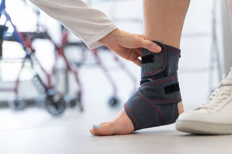 In un ambiente clinico, un operatore sanitario sistema una cavigliera scura al piede di un paziente, con dei deambulatori sullo sfondo.