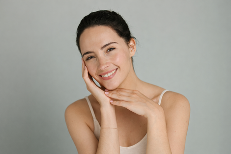 Una persona sorridente con i capelli scuri tirati indietro posa con le mani appoggiate delicatamente sul viso, su uno sfondo grigio.