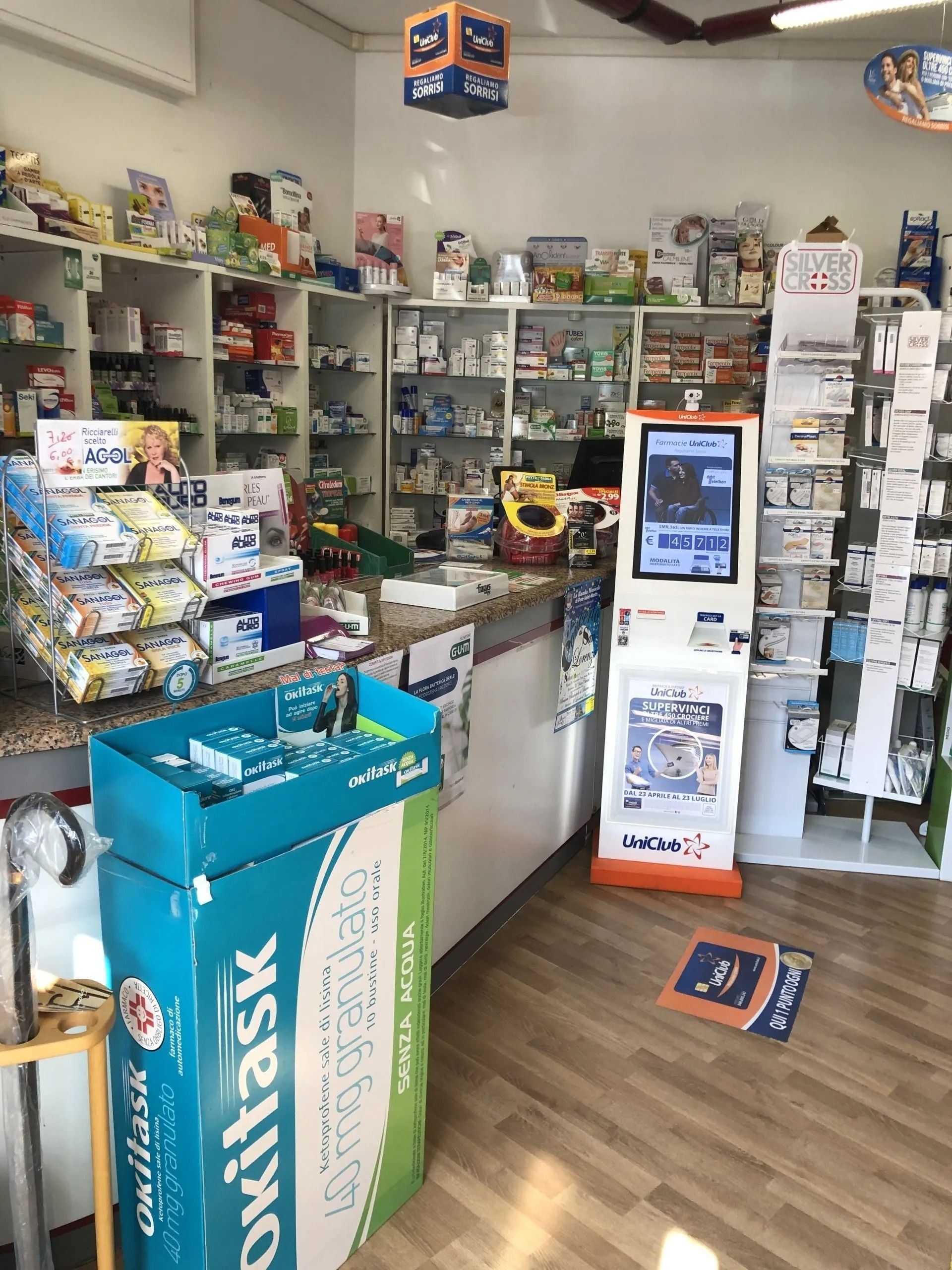 Interno di una farmacia con scaffali pieni di prodotti, un bancone e un chiosco digitale per la salute.