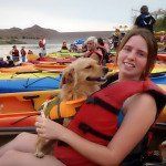 A woman is sitting on a kayak with a dog wearing a life jacket.