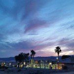 A sunset with palm trees in the foreground and a blue sky with clouds in the background.