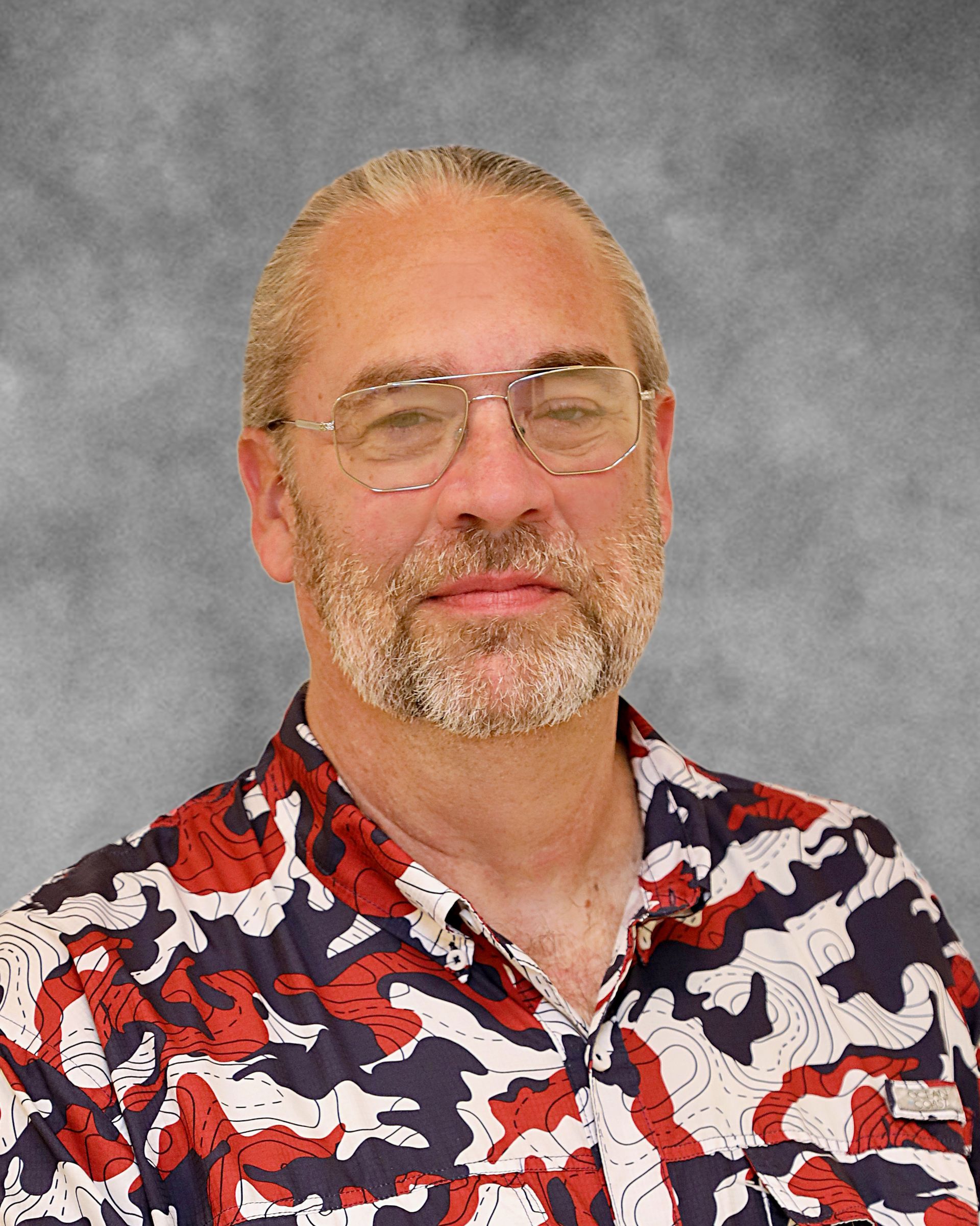 A man with glasses and a beard is wearing a camouflage shirt.