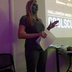 A woman with green paint on her face is giving a presentation in front of a projector screen.