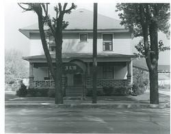 Two-story house with porch, obscured by trees and a pole; street in foreground.