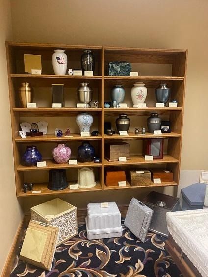 Wooden shelf displaying various cremation urns in a room, with boxes on the floor in front.