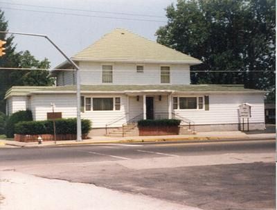 White two-story building with green roof, located at a street corner. Trees and shrubs surround the building.