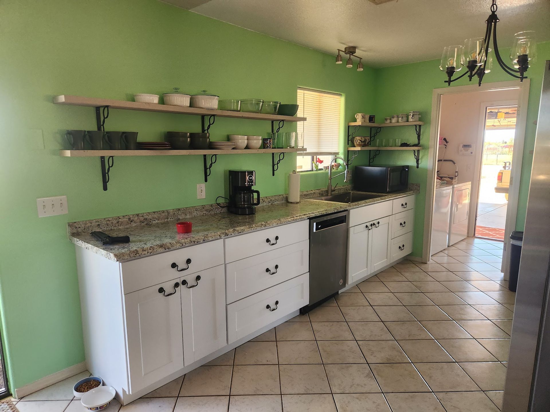 Bright green kitchen with white cabinets, tiled floor, open shelves, and sunlight from the doorway.