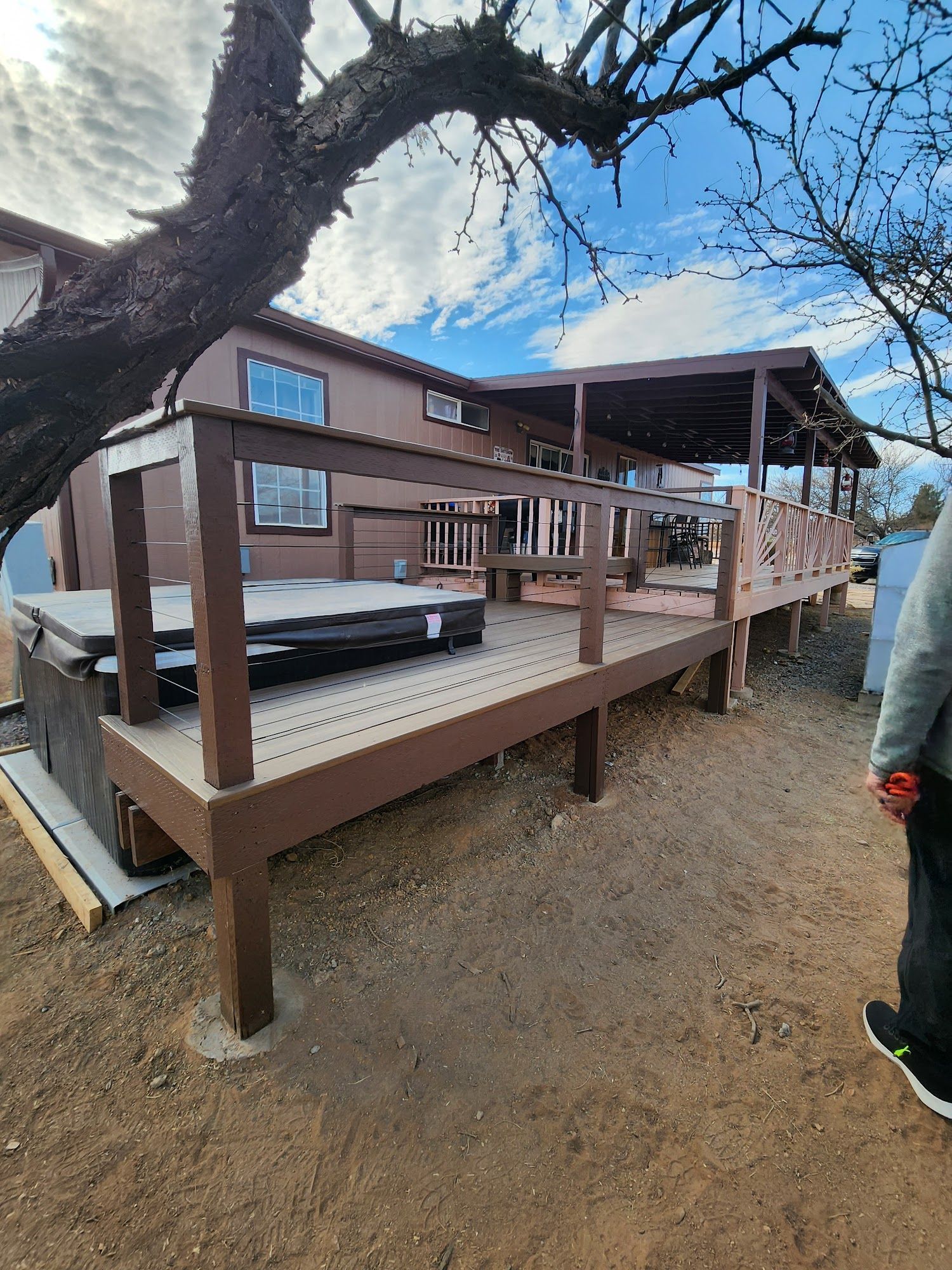 Brown wooden deck with railing beside a house, under a large tree on a dirt yard