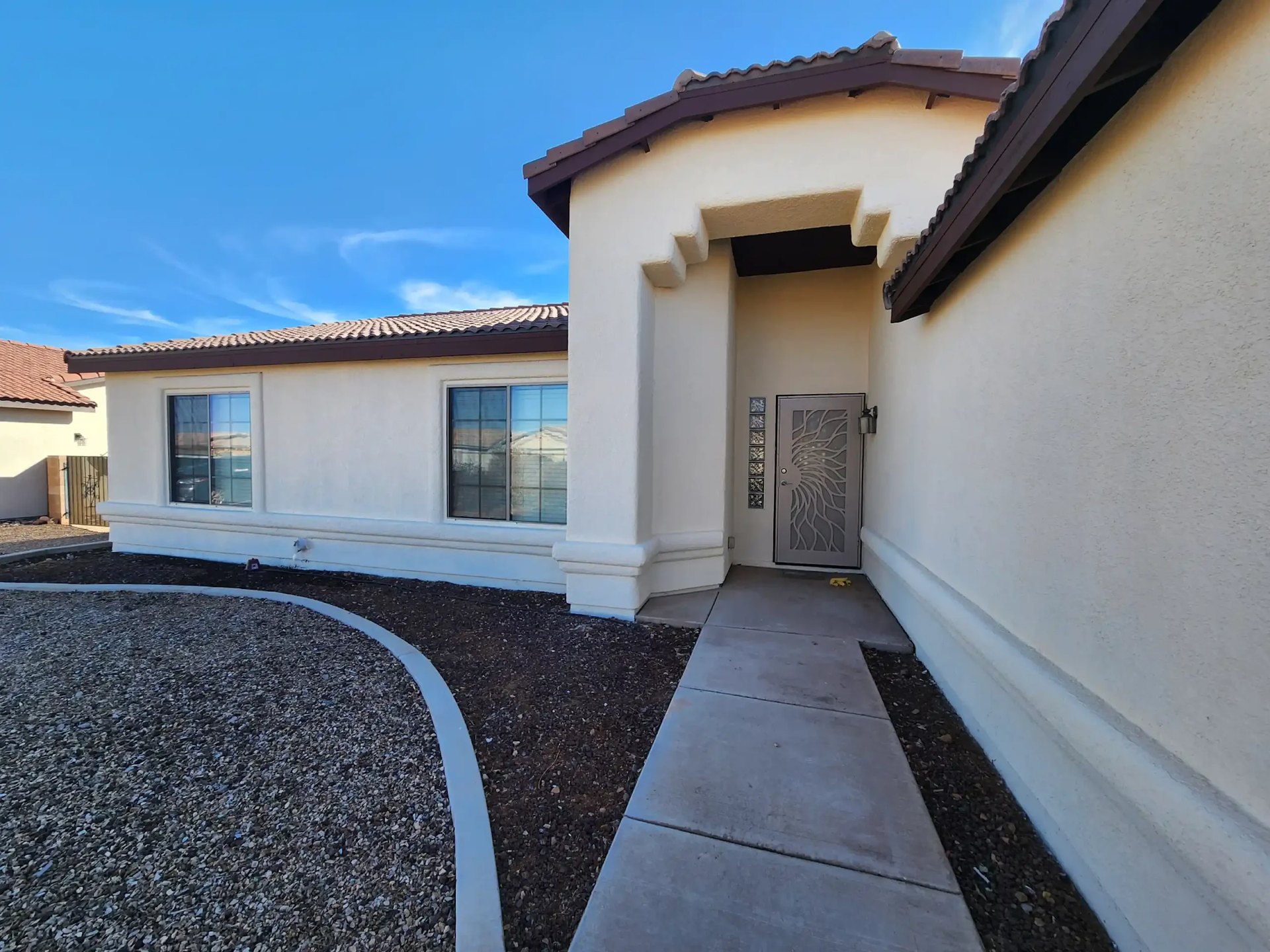 Front walkway to a beige stucco house with white trim and a gravel courtyard under a blue sky