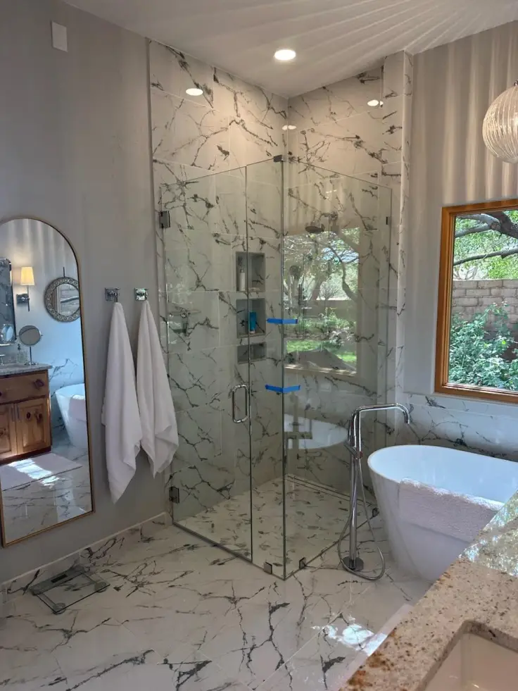 Modern bathroom with glass shower, marble tile, round mirror, tub, and vanity by a window