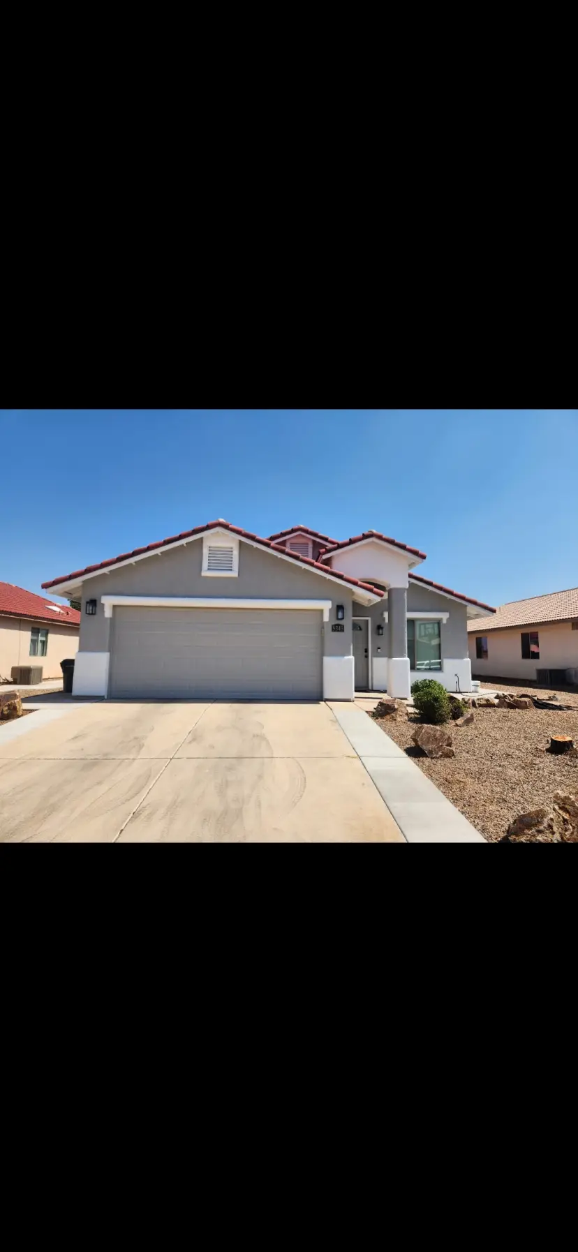 Suburban single-story gray house with a two-car garage and desert landscaping under a clear blue sky