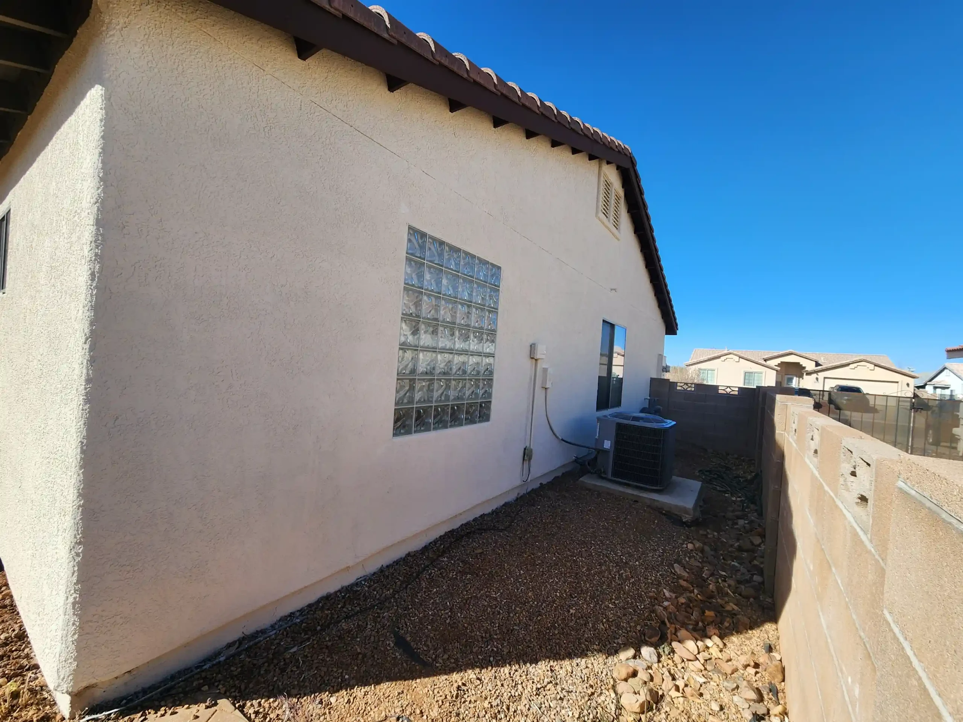 Side yard beside a white stucco house with a glass block window, gravel ground, and blue sky.