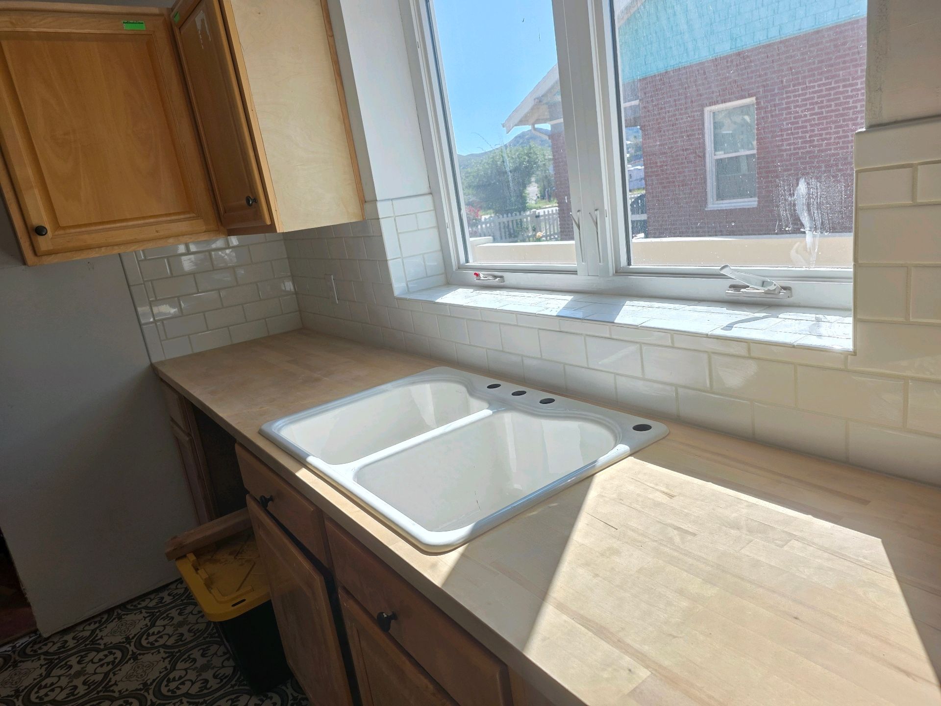 Kitchen countertop with white double sink beneath a window, wood cabinets, and beige tile backsplash.