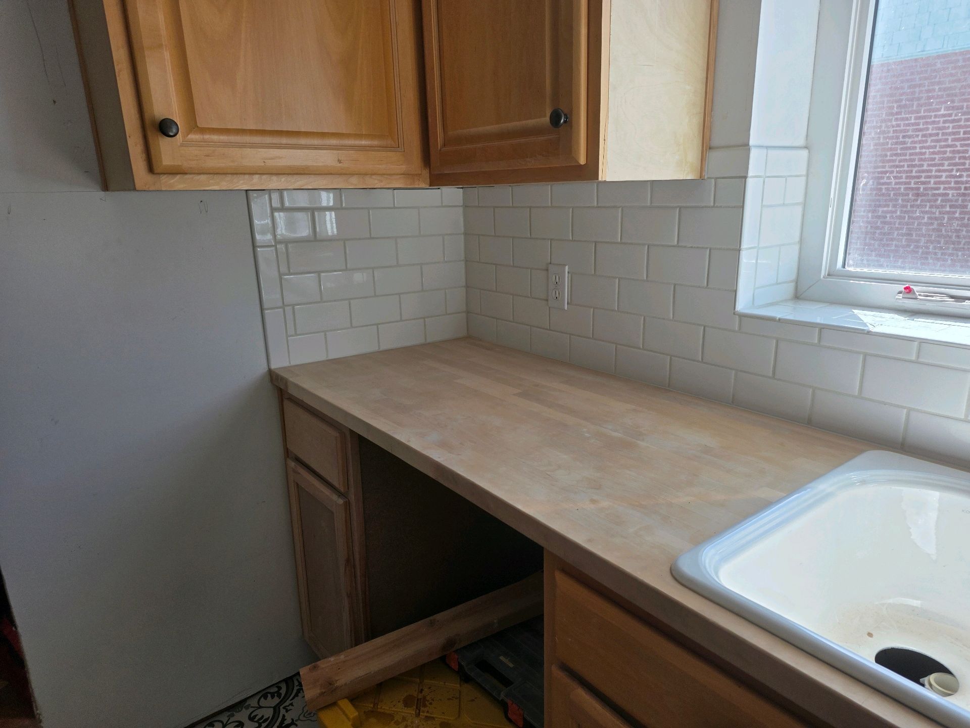 Small kitchen with wooden cabinets, beige countertop, white tile backsplash, and a sink by a window.