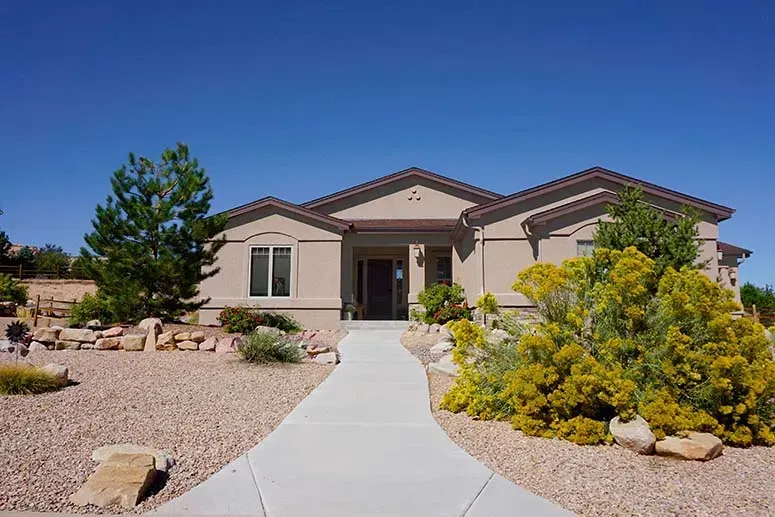 Single-story beige house with a front walkway, gravel landscaping, and yellow shrubs under a clear blue sky