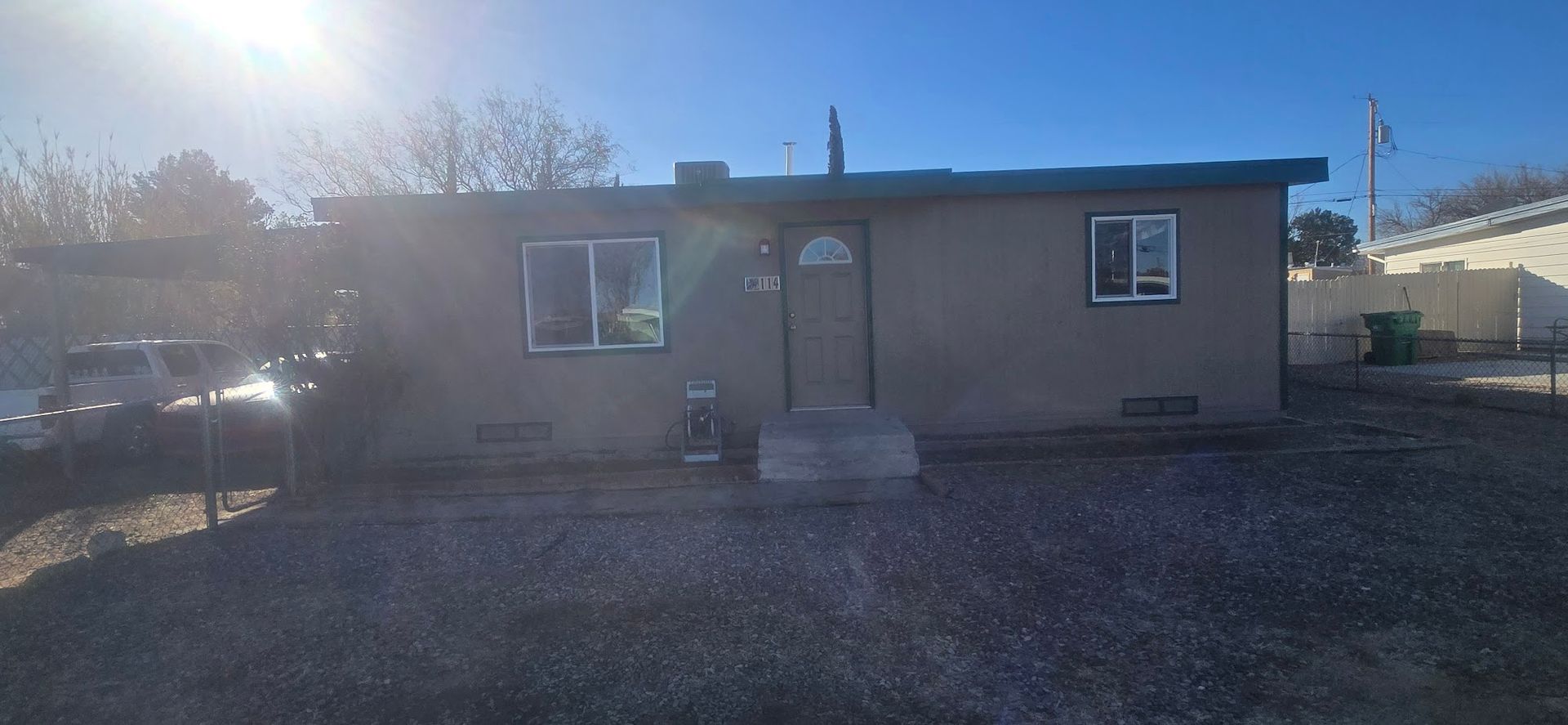 Small beige house with a front door and windows, seen in bright sunlight on a gravel lot.