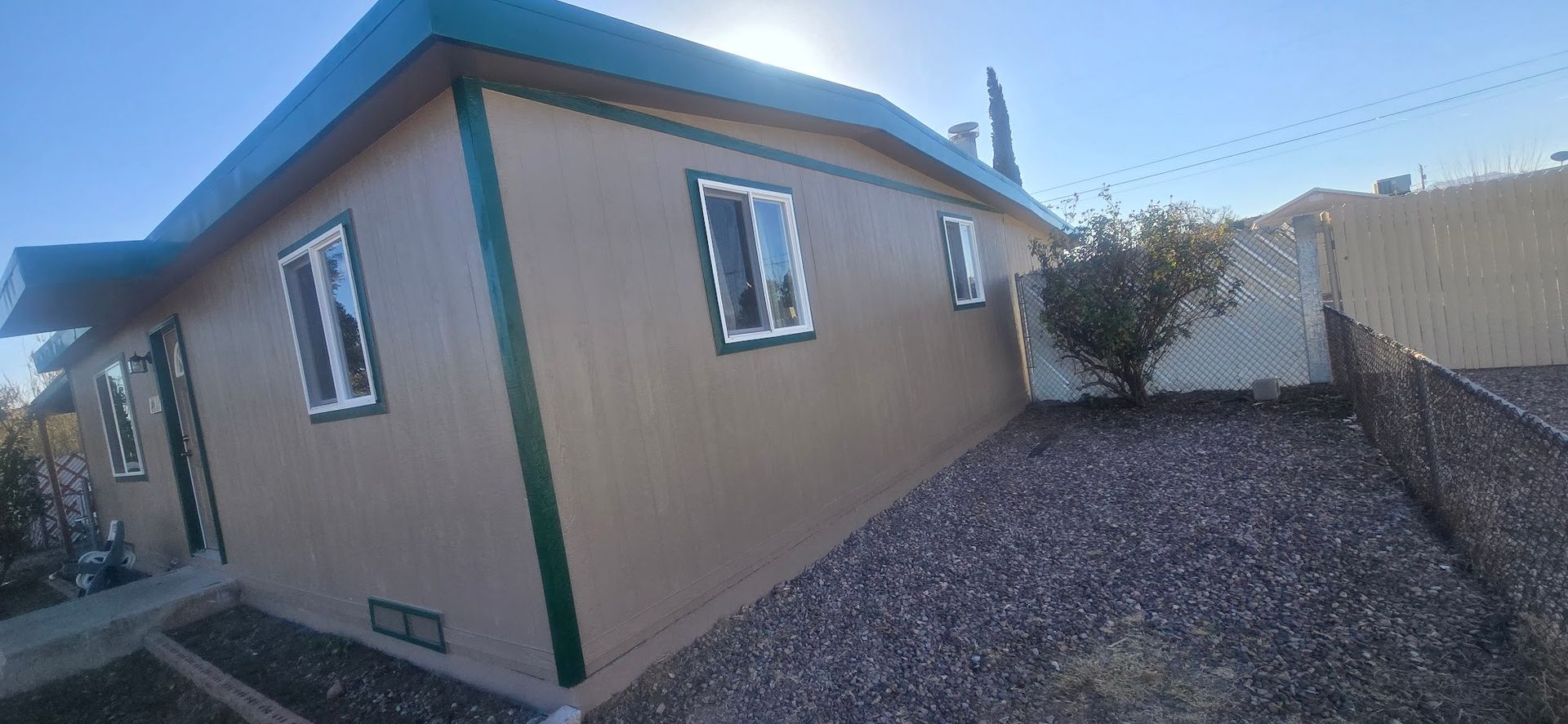 Tan stucco house with green trim beside a gravel yard under a blue sky.