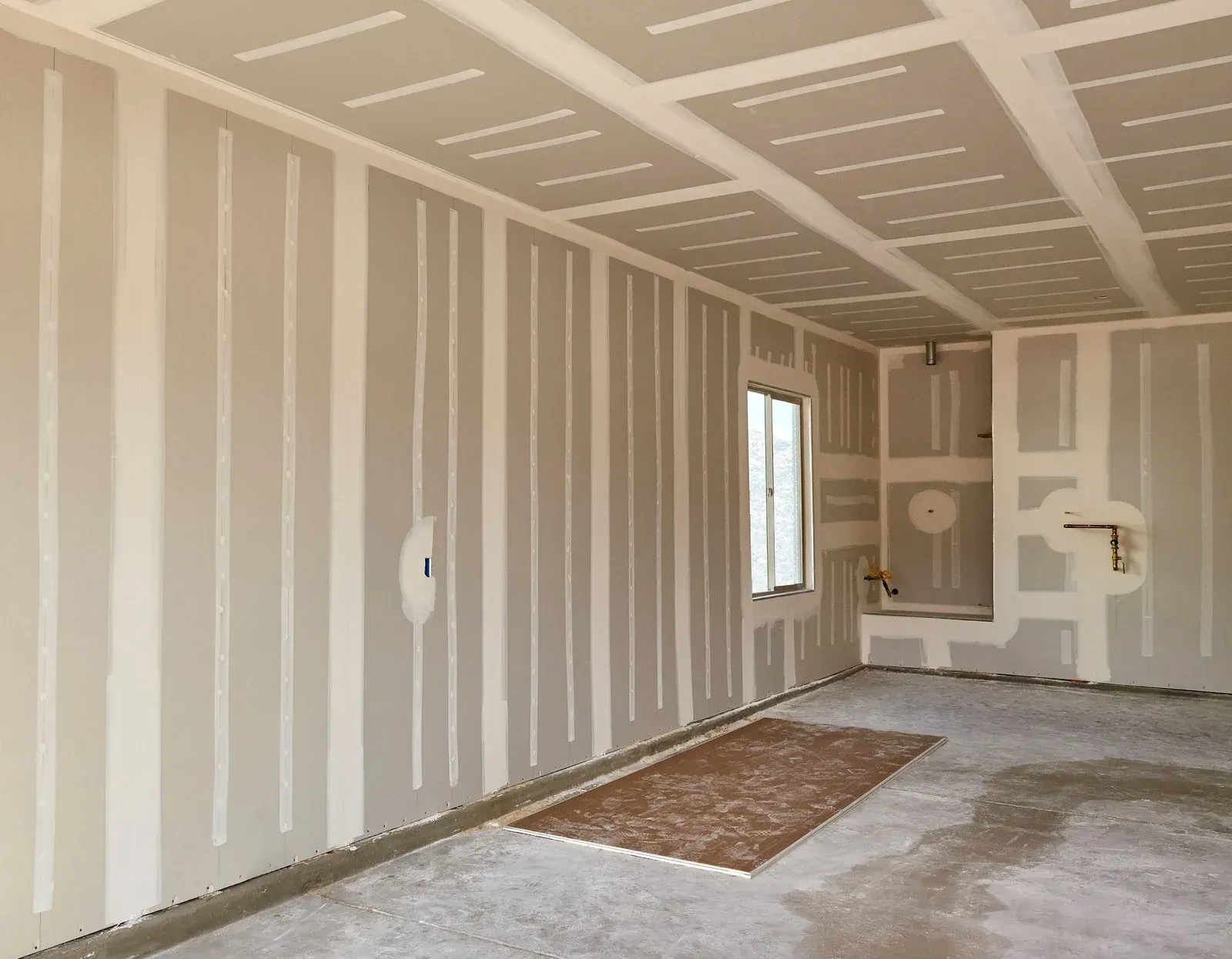 Unfinished drywall room with white walls, exposed seams, and a small window