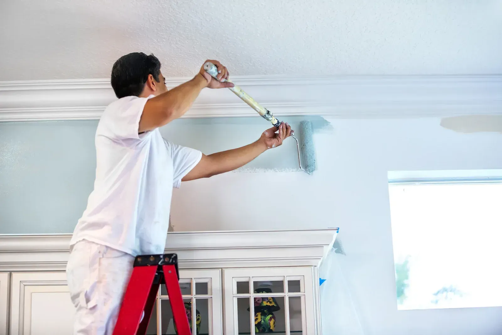 Man painting a white ceiling with a roller while standing on a red ladder in a bright room