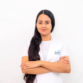 Uma mulher de cabelos longos está vestindo um jaleco branco e está de pé com os braços cruzados.
