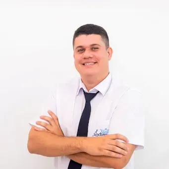 Um homem de camisa branca e gravata está de pé com os braços cruzados e sorrindo.