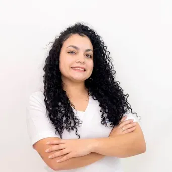 Uma mulher com cabelos cacheados está de pé com os braços cruzados e sorrindo.