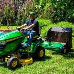 Man on green riding lawnmower with a towed leaf collector on a lawn.