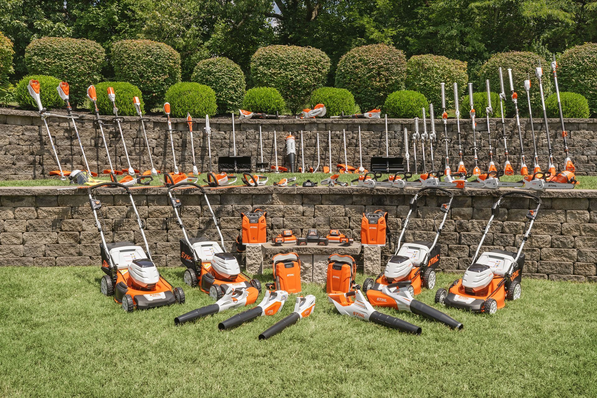 A collection of orange and white STIHL outdoor power tools arranged on grass, in front of a stone wall and hedges.