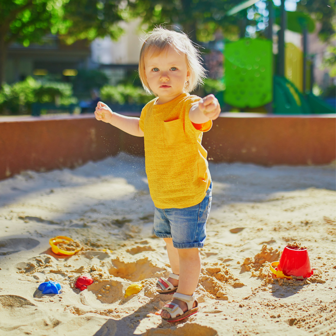A little girl in a yellow shirt is standing in a sandbox.