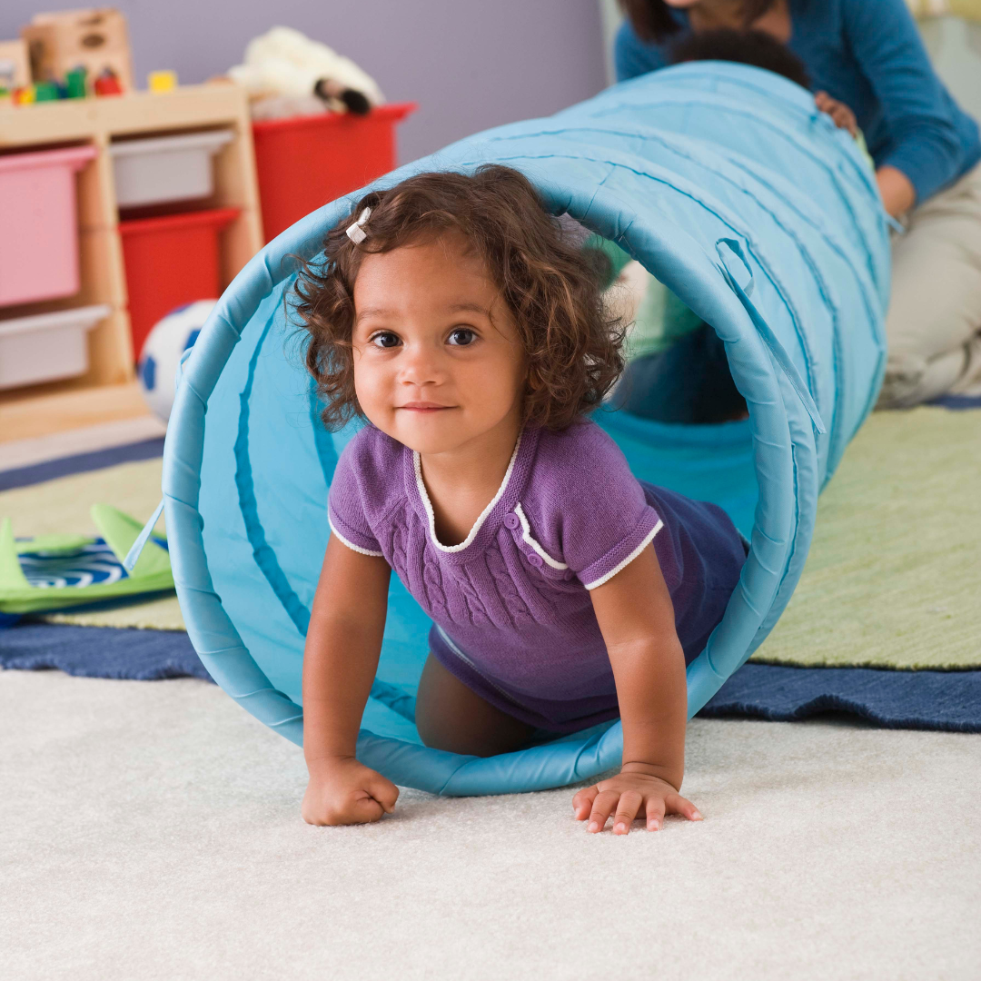 A little girl in a purple shirt is crawling through a blue tunnel