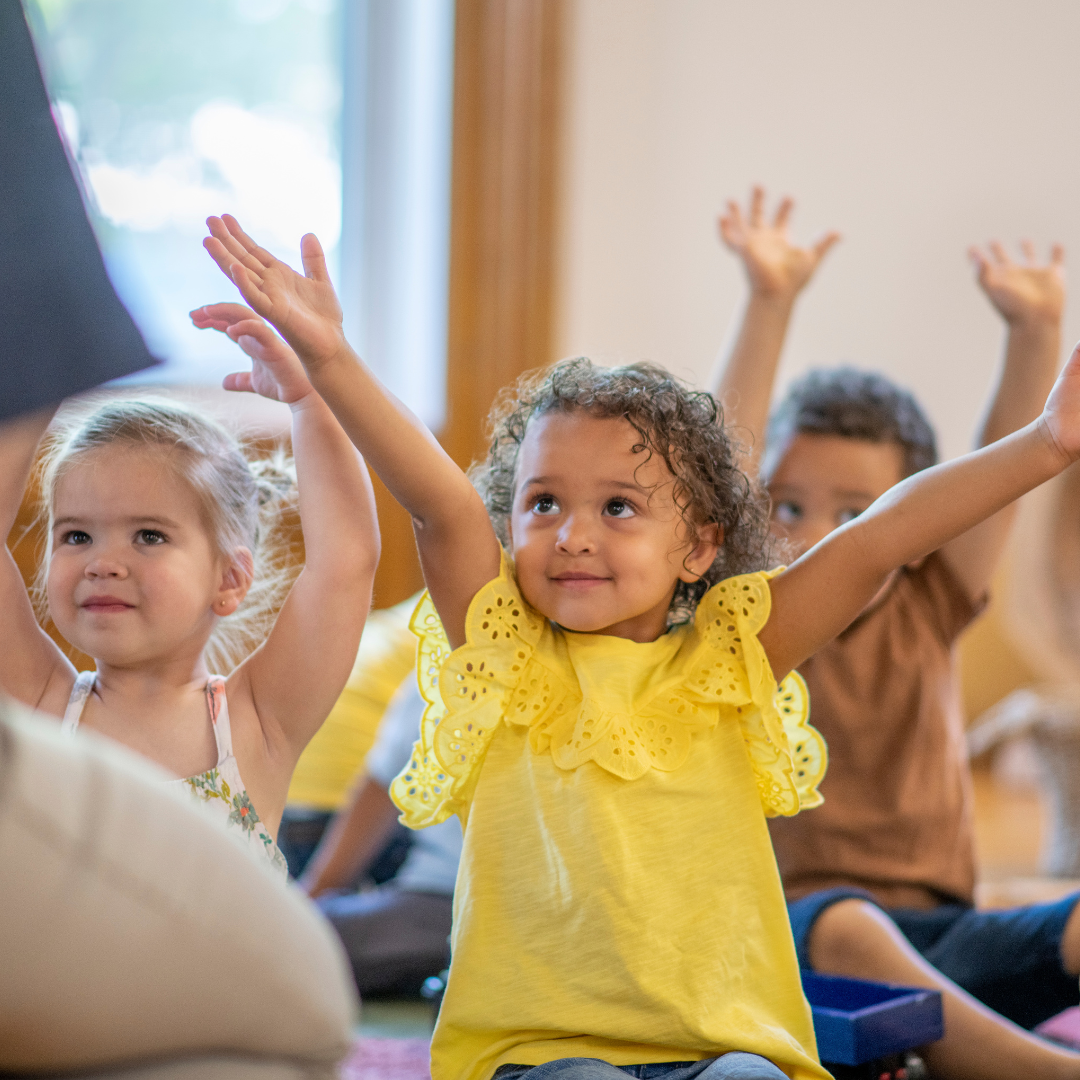 A group of children are sitting on the floor with their arms in the air.