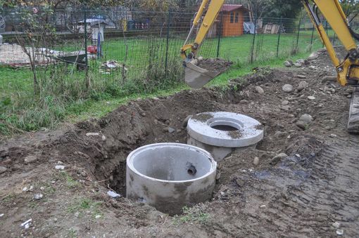 A yellow excavator is digging a hole in the dirt next to two concrete rings.
