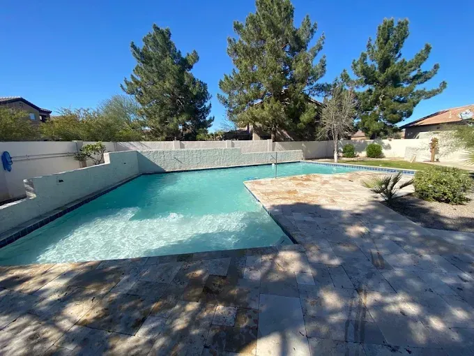 A turquoise pool in a backyard with stone patio, trees, and clear blue sky.