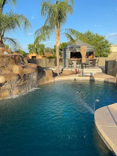 Pool with waterfall feature and outdoor bar under a palm tree in a sunny setting.