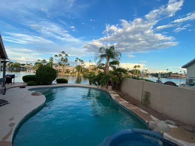 Swimming pool overlooking a lake with blue sky and clouds. Palm trees and homes are visible.