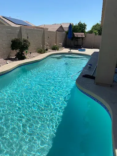 Blue swimming pool in a sunny backyard, surrounded by a stone patio and walls.