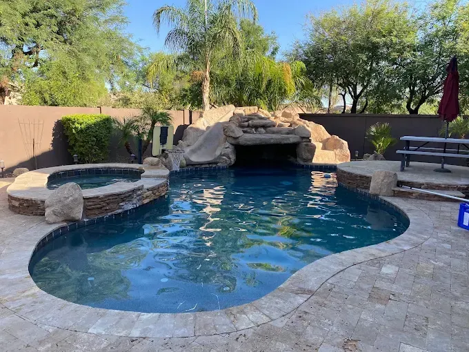Backyard pool with spa, waterfall, and slide, surrounded by stone and lush greenery under a bright blue sky.