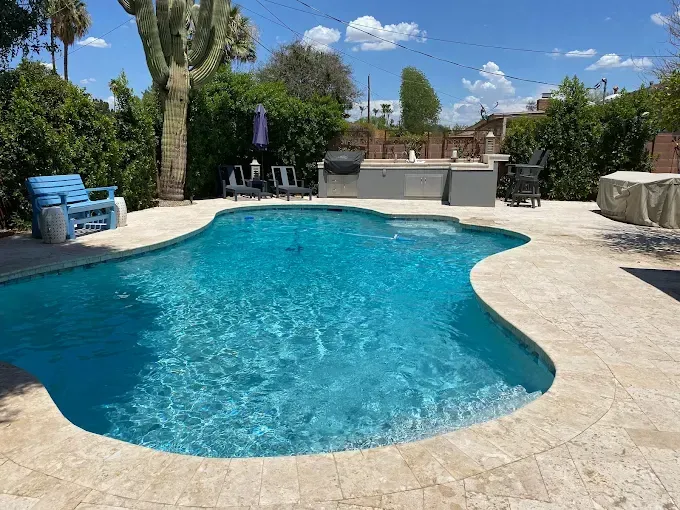 Pool in backyard with built-in grill and seating. Bright blue water, beige stone surround, and green trees.