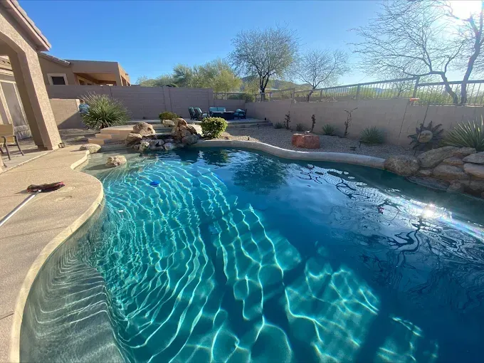 A backyard swimming pool with clear blue water and desert landscaping, under a sunny sky.