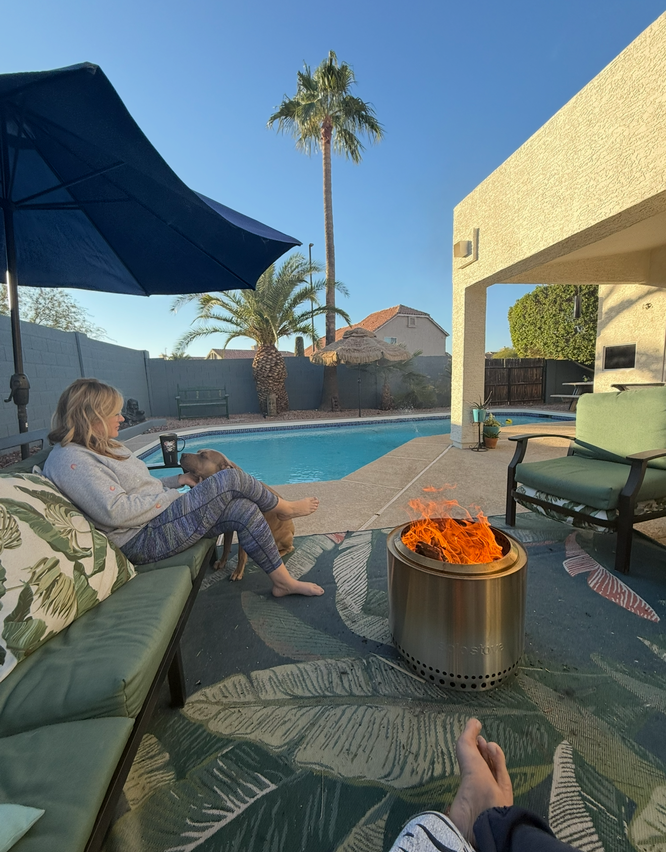 Woman relaxing by a pool with a fire pit, dog, and patio furniture on a sunny day.