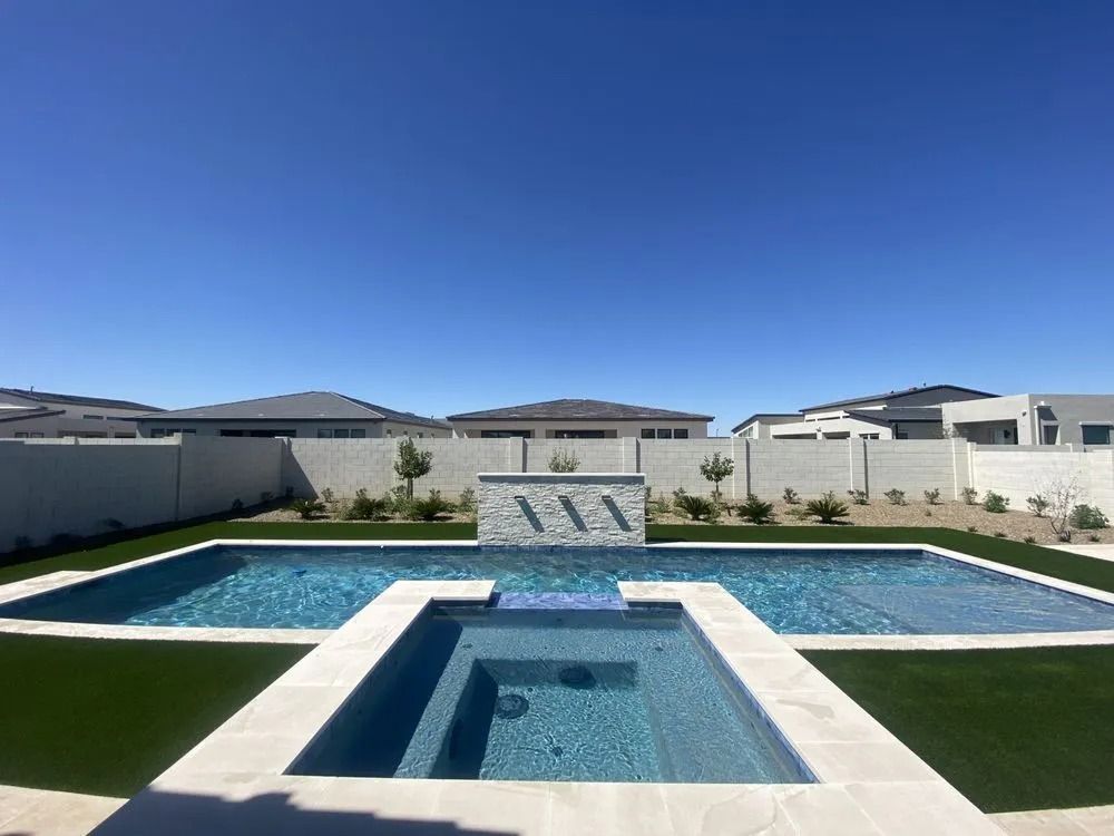 Pool and spa in a backyard with a water feature, artificial turf, and a clear blue sky.